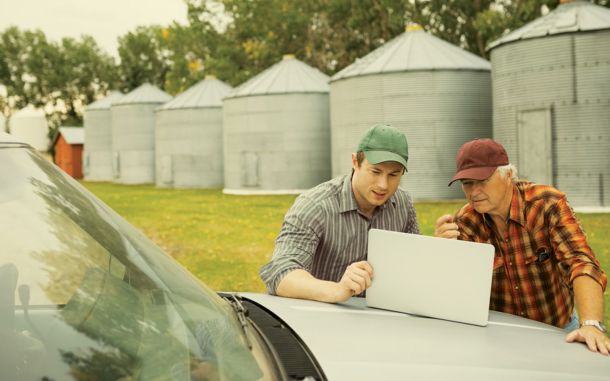 Two farmers looking at a laptop on the hood of a pickup while standing outside in front of aluminum grain bins