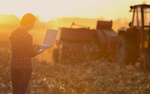 Woman standing in field with laptop with tractor in the background.